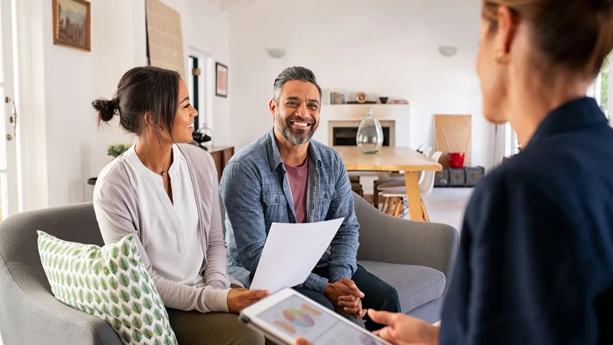 A smiling couple on a sofa reviewing papers with a Realtor who holds a tablet in a bright living space